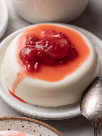 Panna cotta topped with homemade strawberry sauce on a speckled white plate. A spoon rests next to it.