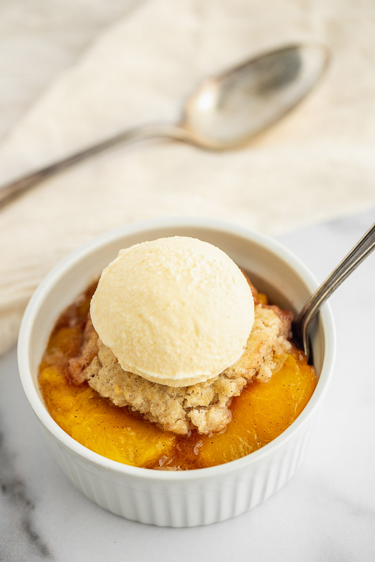 A white ramekin on a marble surface filled with peach cobbler and a scoop of ice cream on top.