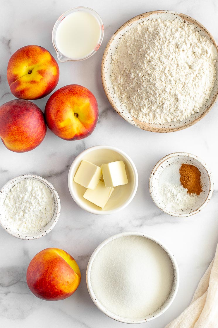 The ingredients for peach cobbler in various bowls and plates on a marble surface.