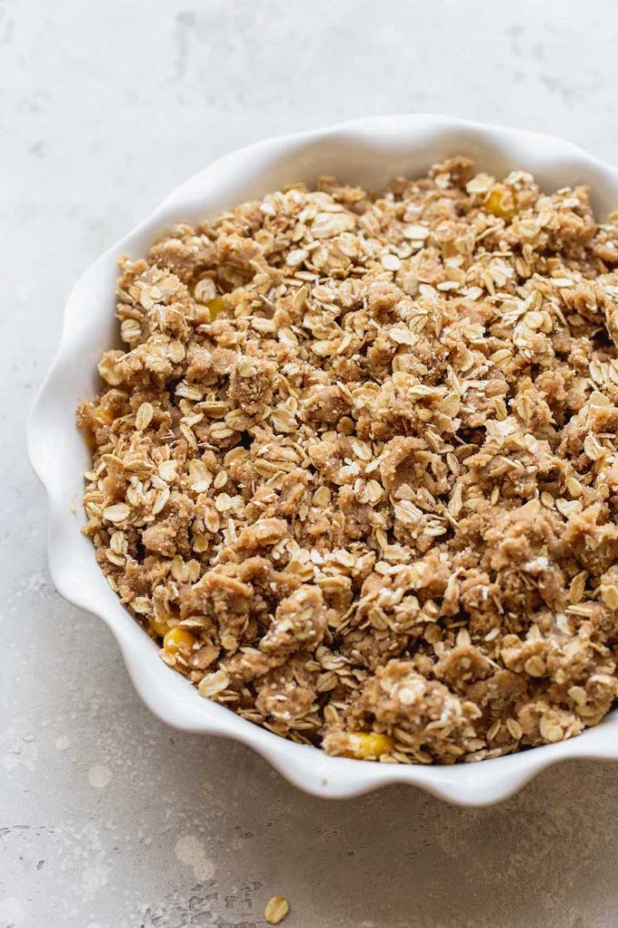 A round white baking dish with sliced peaches topped with the oat topping ready to be baked.