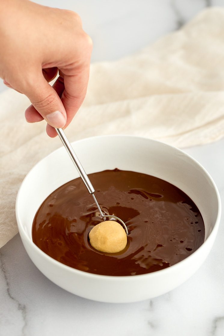 A white bowl filled with melted chocolate and a ball of the peanut butter mixture being dipped in it. 