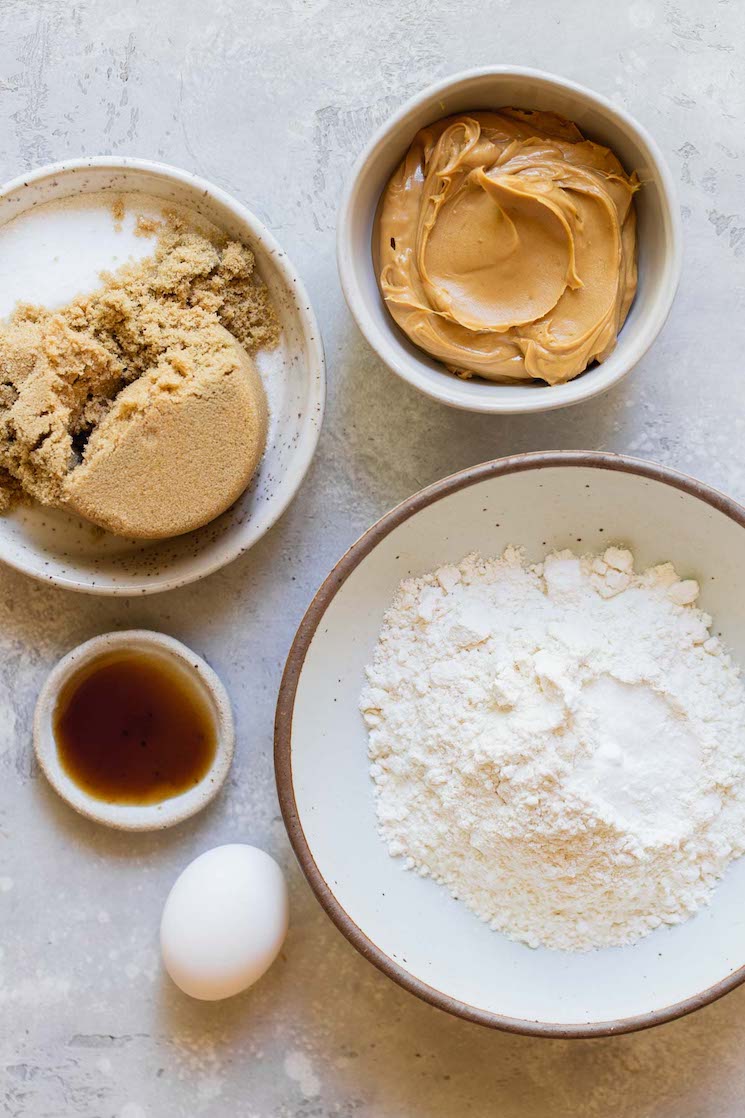 The ingredients needed to make peanut butter cookies in bowls on top of a rustic gray surface.