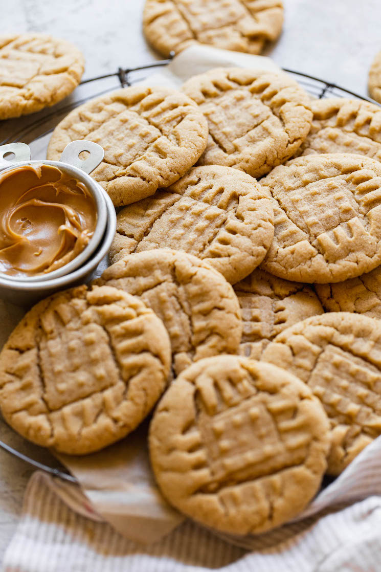 A round cooling rack holding peanut butter cookies piled on each other with a small metal cup of peanut butter off to the side.
