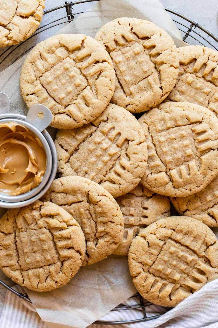 An overhead view of peanut butter cookies on a round cooling rack with a metal cup of peanut butter off to the side.