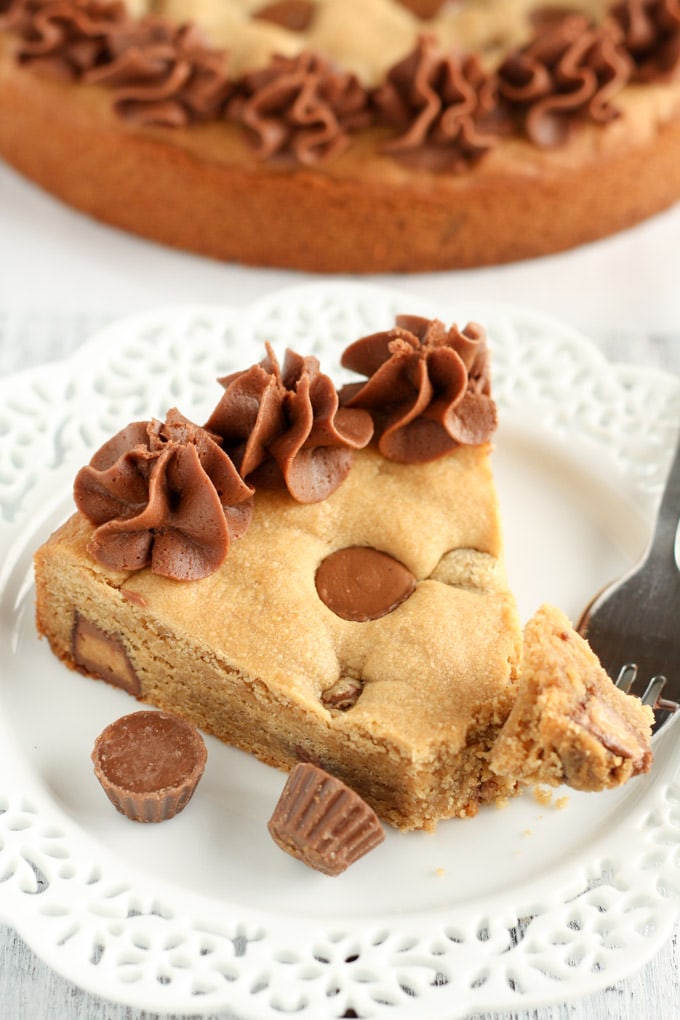 A slice of homemade cookie cake on a white plate with a fork. The rest of the cookie cake rests in the background. 