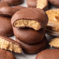 A stack of peanut butter eggs. The top one has a bite missing and a small bowl of peanut butter and more chocolate coated eggs surround it.