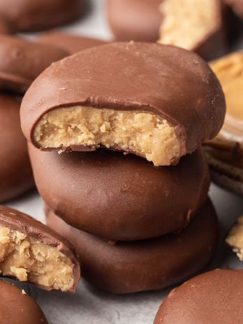 A stack of peanut butter eggs. The top one has a bite missing and a small bowl of peanut butter and more chocolate coated eggs surround it.