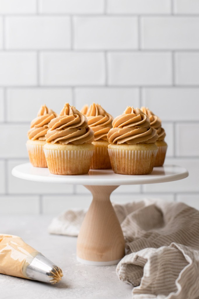 A marble cake stand holding vanilla cupcakes topped with peanut butter frosting and a piping bag of frosting off to the side.