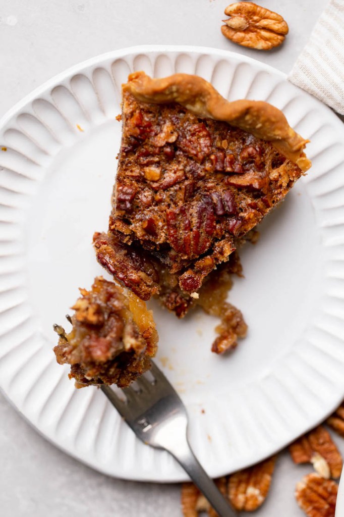 An individual slice of pecan pie on a white plate with a bite taken out on a fork.