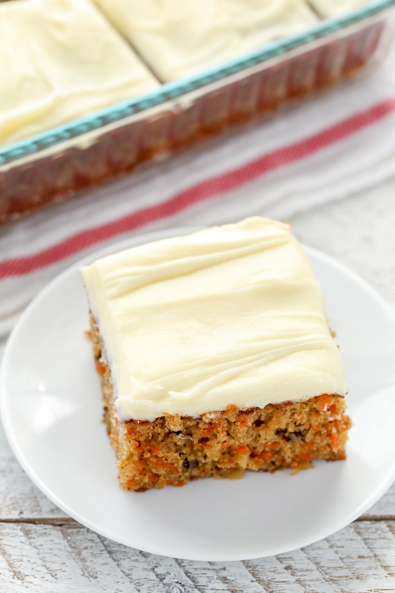 A slice of pineapple carrot cake on a white plate. A pan of cake rests in the background. 