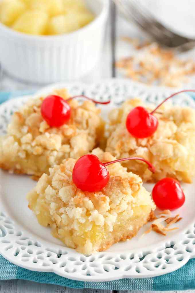 Three pineapple bars garnished with maraschino cherries on a white dessert plate. A small bowl of pineapple rests in the background.