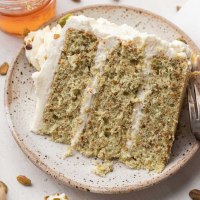A slice of pistachio cake on a speckled plate. A small jar of honey and pistachios are near the plate.