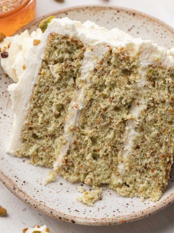 A slice of pistachio cake on a speckled plate. A small jar of honey and pistachios are near the plate.