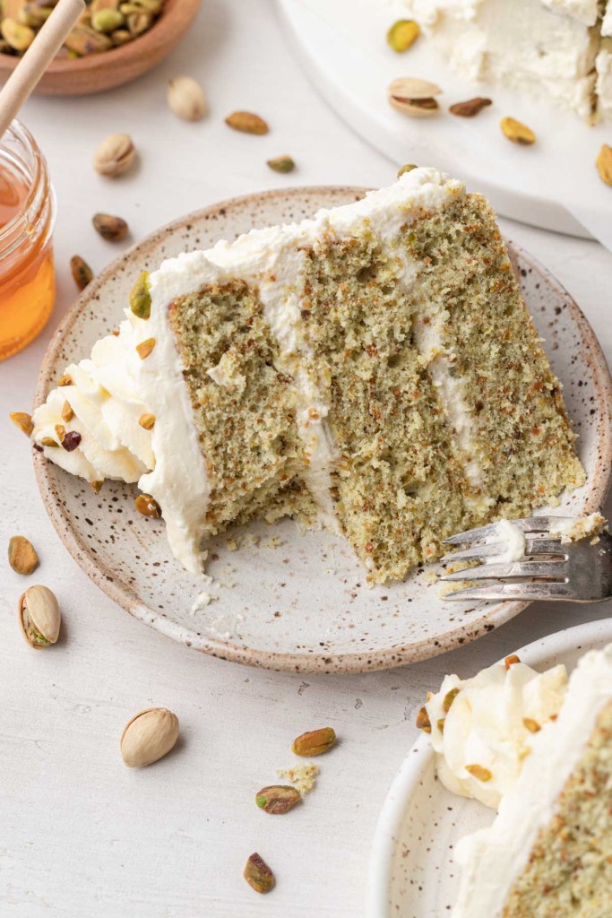 A slice of pistachio layer cake on a speckled white dessert plate, with a bite missing. 