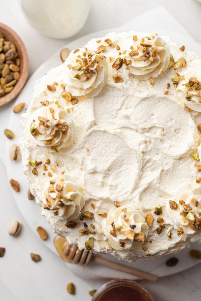 An overhead view of a frosted pistachio cake, with pistachios and honey on the side. 