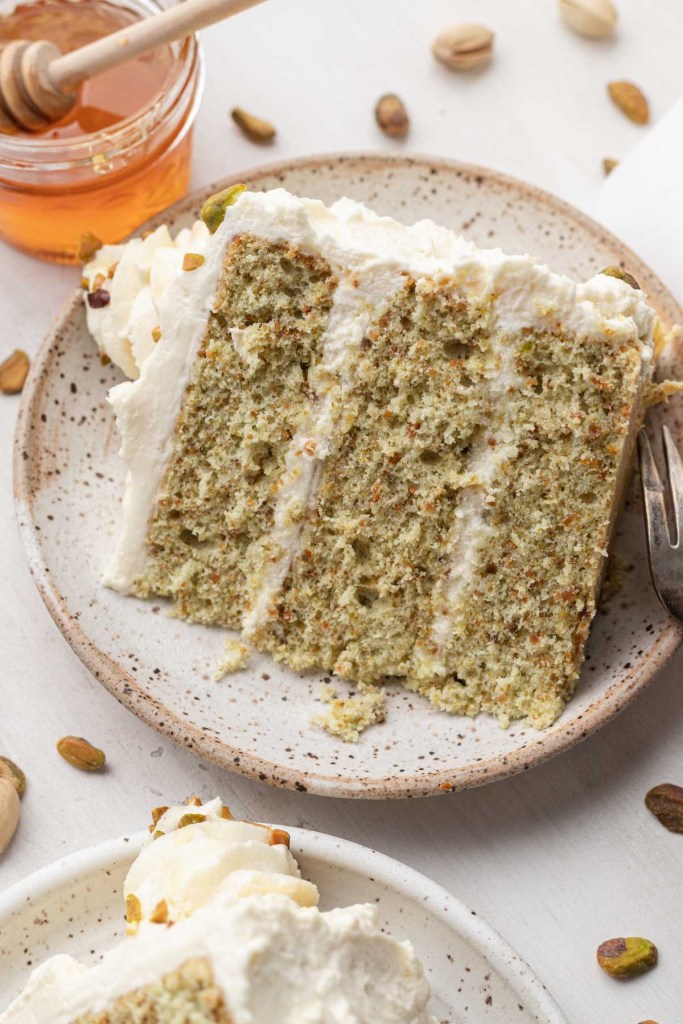 A slice of pistachio nut cake on a speckled white dessert plate, with honey on the side. 