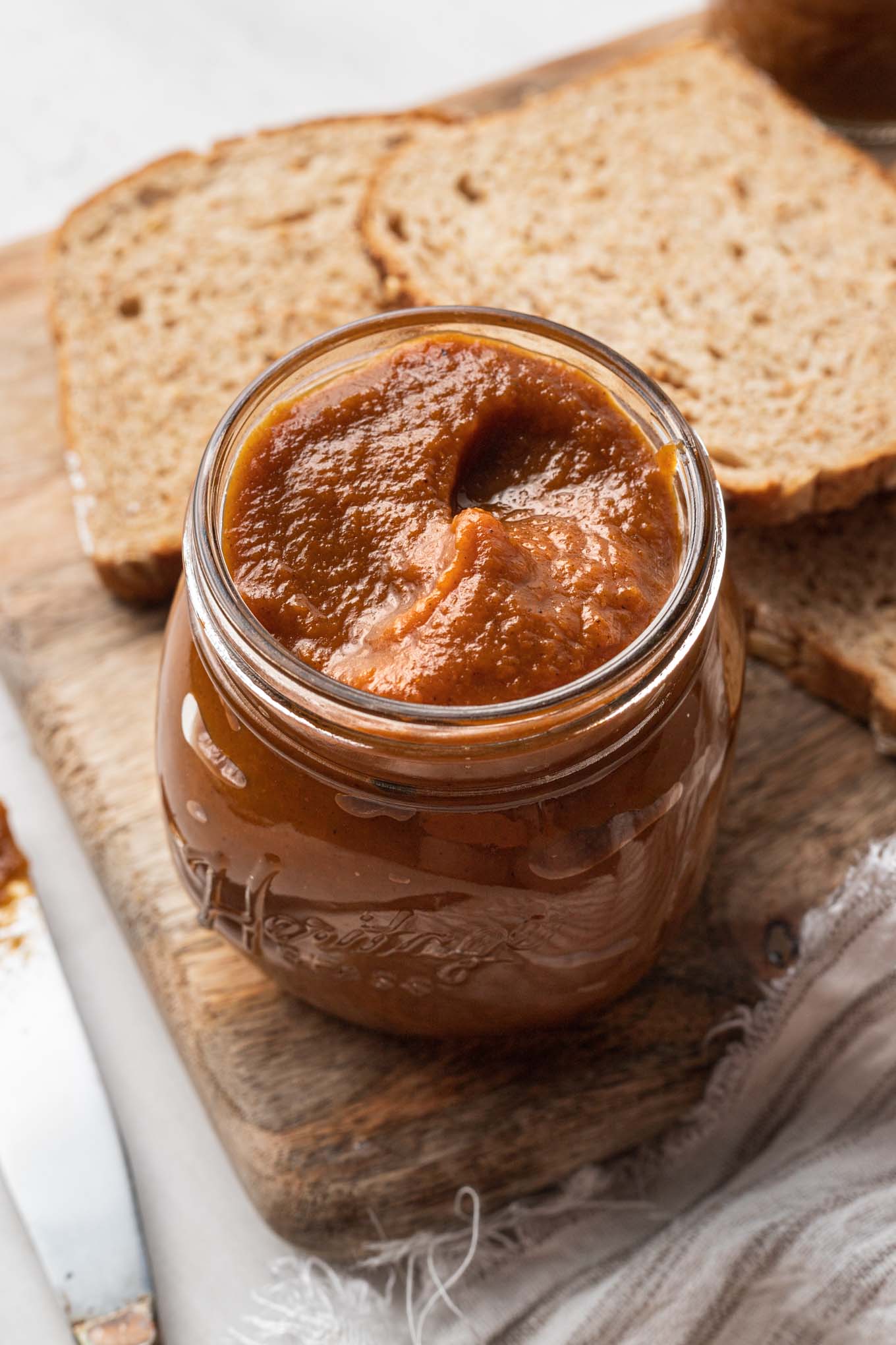 A jar of pumpkin butter on a wooden cutting board with two pieces of bread. 