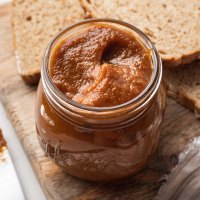 A jar filled with pumpkin butter on a wooden cutting board. A couple slices of bread sit behind the jar.