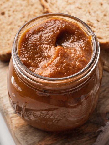 A jar filled with pumpkin butter on a wooden cutting board. A couple slices of bread sit behind the jar.