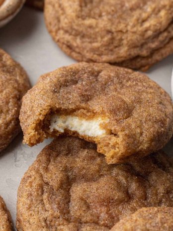 Pumpkin cheesecake cookies on a gray surface. One cookie has a bite missing to show the cream cheese center.