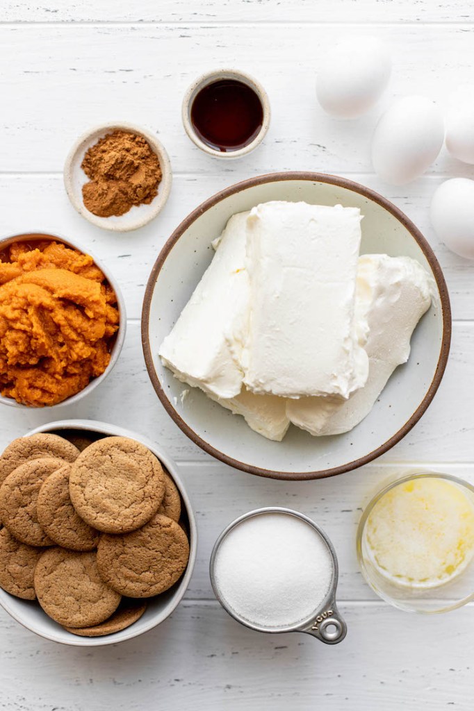 The ingredients needed to make a pumpkin cheesecake sitting on top of on a white wood surface.