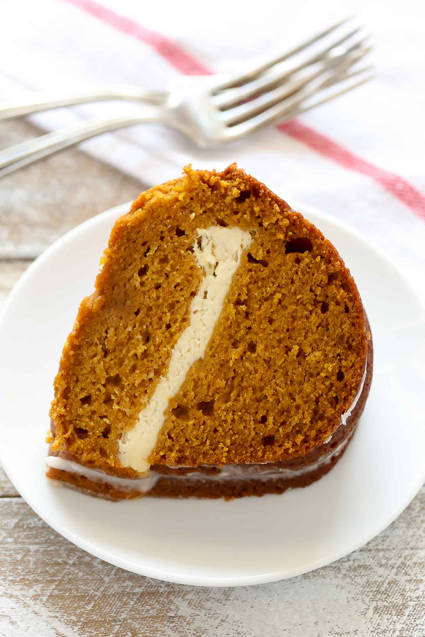 A slice of pumpkin cream cheese cake on a white plate. Two forks and a tea towel rest in the background. 