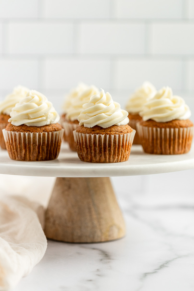 A group of pumpkin cupcakes topped with cream cheese frosting setting on top of a marble cake stand.