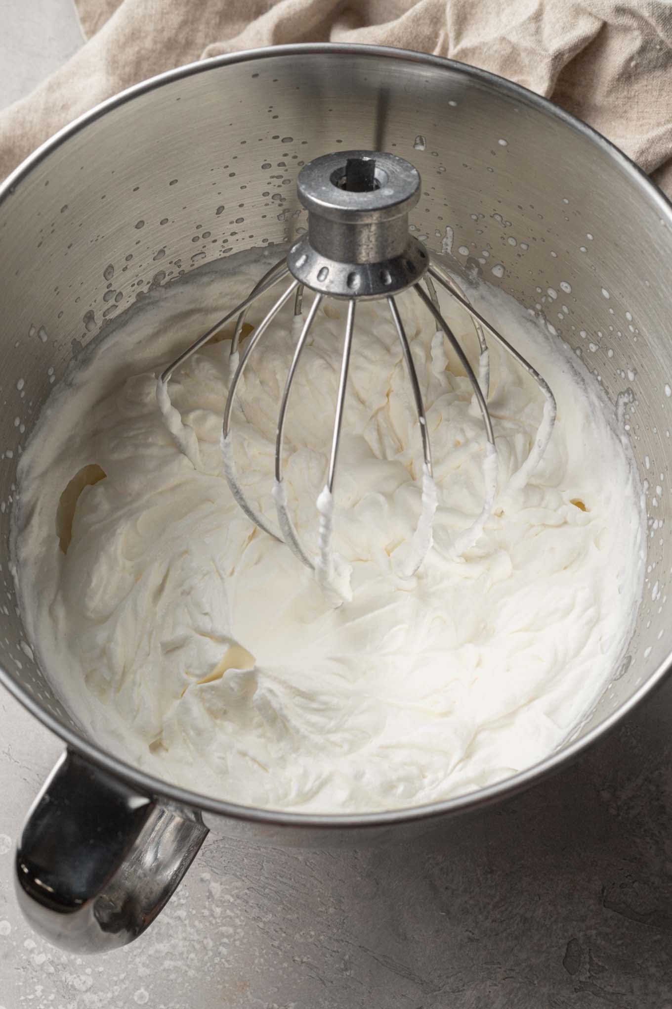 An overhead view of whipped cream in the bowl of a stand mixer, with a whisk attachment. 