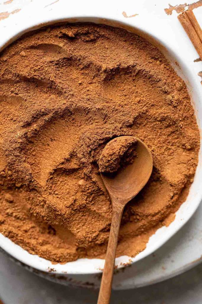 Pumpkin pie spice mixed together in a white dish. A wooden spoon rests on top of the spices and a cinnamon stick rests on the side.