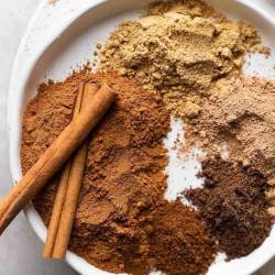 Several different spices separated on a white plate. Two cinnamon sticks rest on top of the ground cinnamon.
