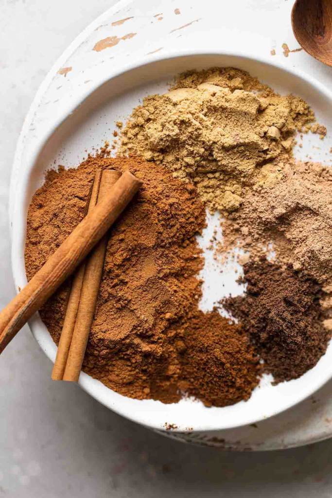 Several different spices separated on a white plate. Two cinnamon sticks rest on top of the ground cinnamon.