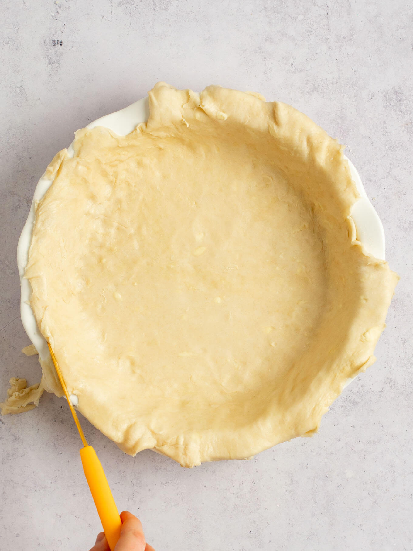 An overhead view of pie dough in a pie dish. A knife is being used to trim the edges.