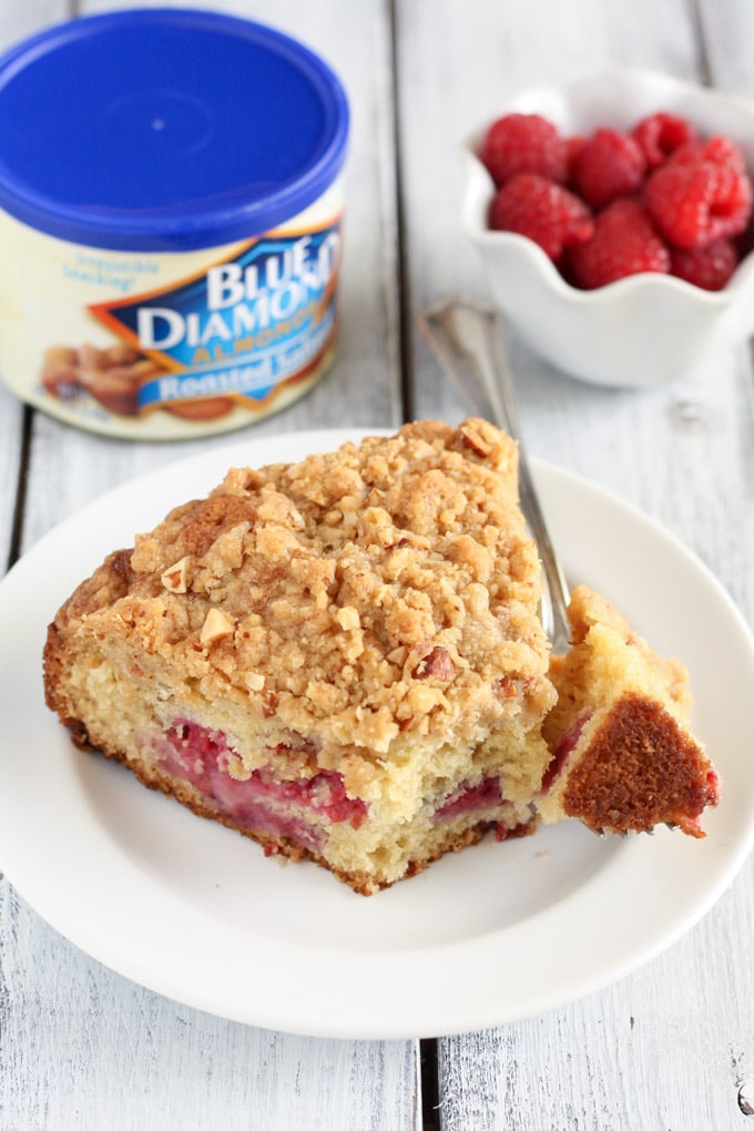 A slice of raspberry crumb cake on a white plate. A bowl of fresh raspberries and a container of almonds rest in the background. 