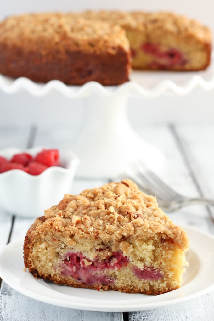 A slice of raspberry almond cake with crumb topping on a white plate. A dish of fresh raspberries and a cake on a cake stand rest in the background. 