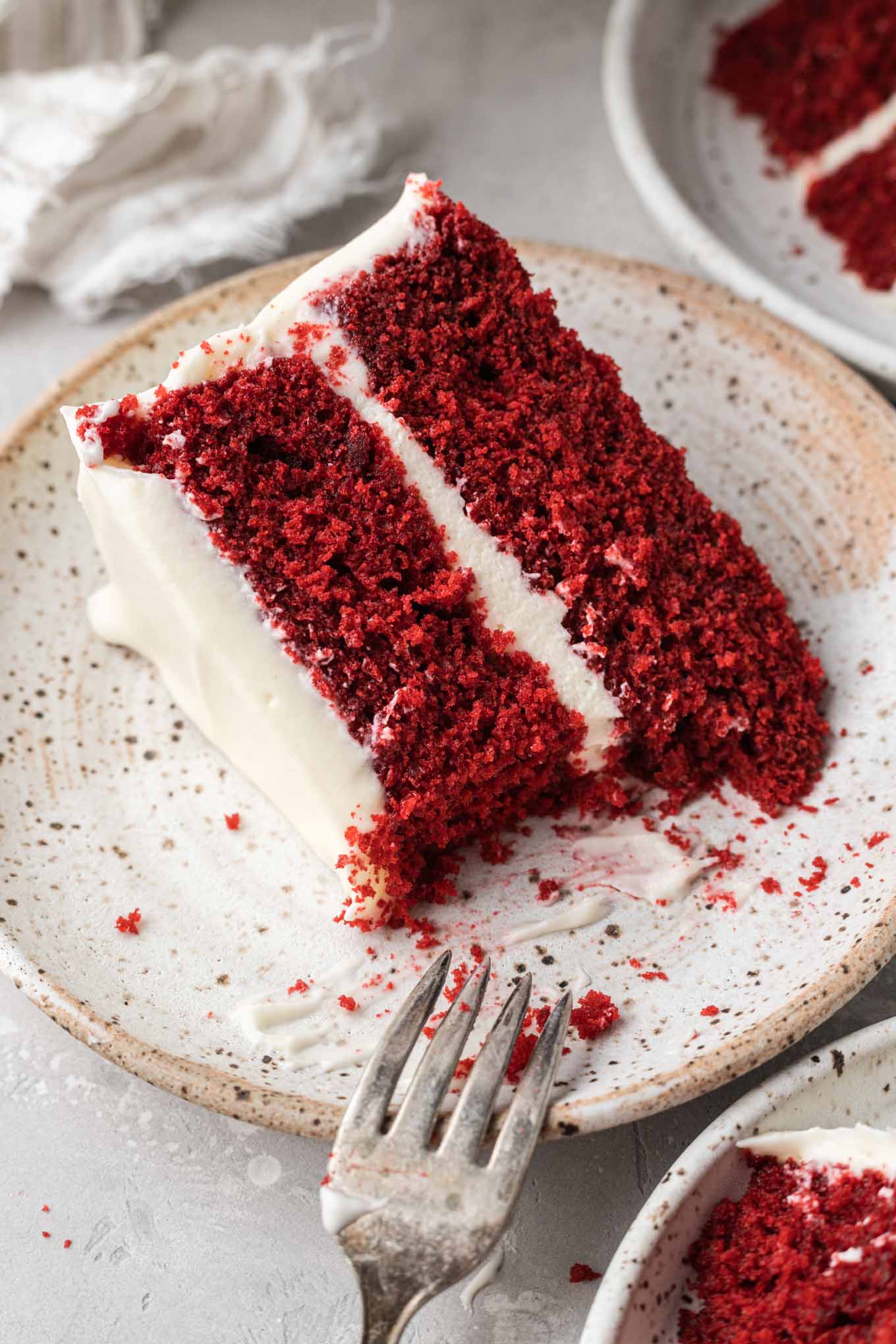 A slice of frosted red velvet cake on a speckled white plate. 