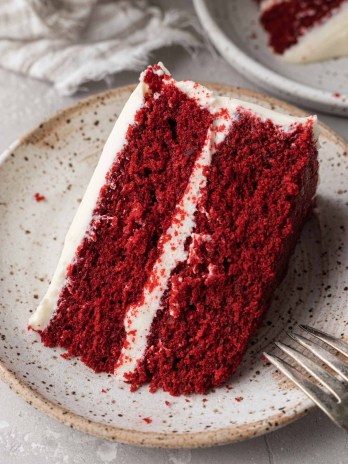 A slice of red velvet cake on a speckled white plate. A fork rests on the plate.