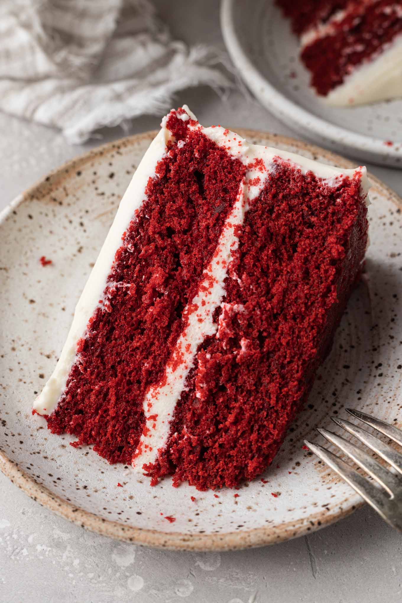 A slice of frosted red velvet cake on a speckled dessert plate. 