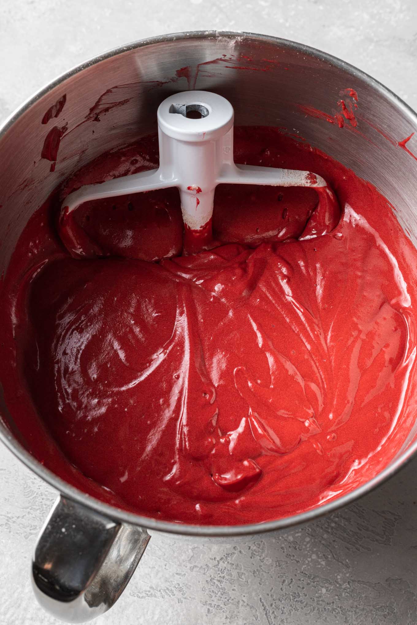 An overhead view of red velvet cake batter in a mixing bowl with a paddle attachment.
