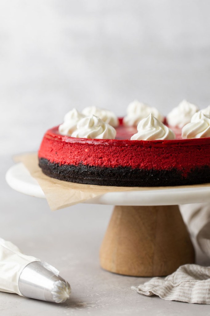 Side view of a red velvet cheesecake topped with whipped cream on a cake stand. A piping bag rests on the side. 