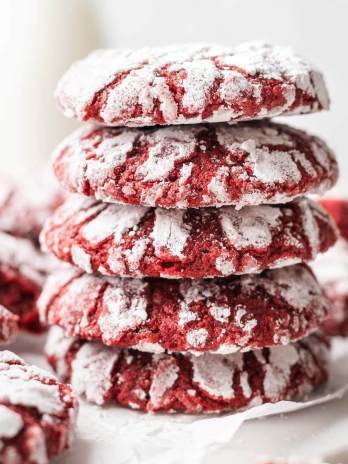 A stack of four red velvet crinkle cookies. More cookies and a small jug of milk are in the background.