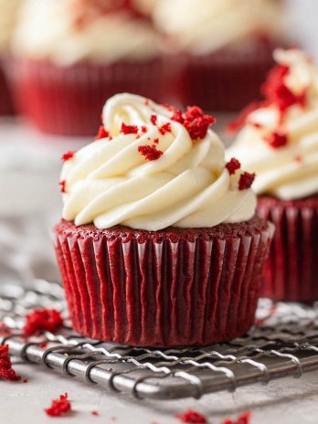 Two red velvet cupcakes topped with cream cheese frosting on a wire rack.