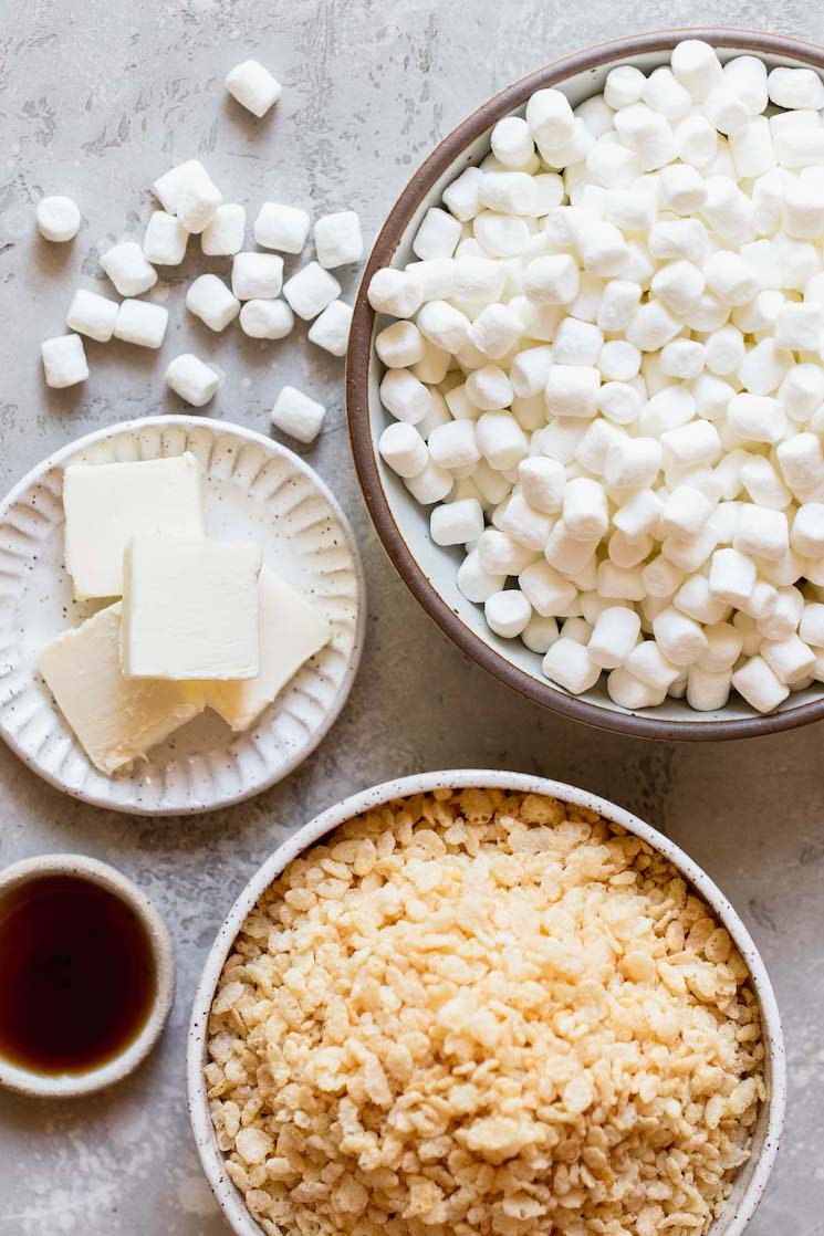 Marshmallows, sliced butter, vanilla extract, and cereal laying out on a gray surface in white and brown plates.