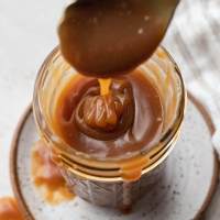 An overhead view of salted caramel sauce in a jar. A spoon is being lifted from the jar.