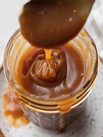 An overhead view of salted caramel sauce in a jar. A spoon is being lifted from the jar.