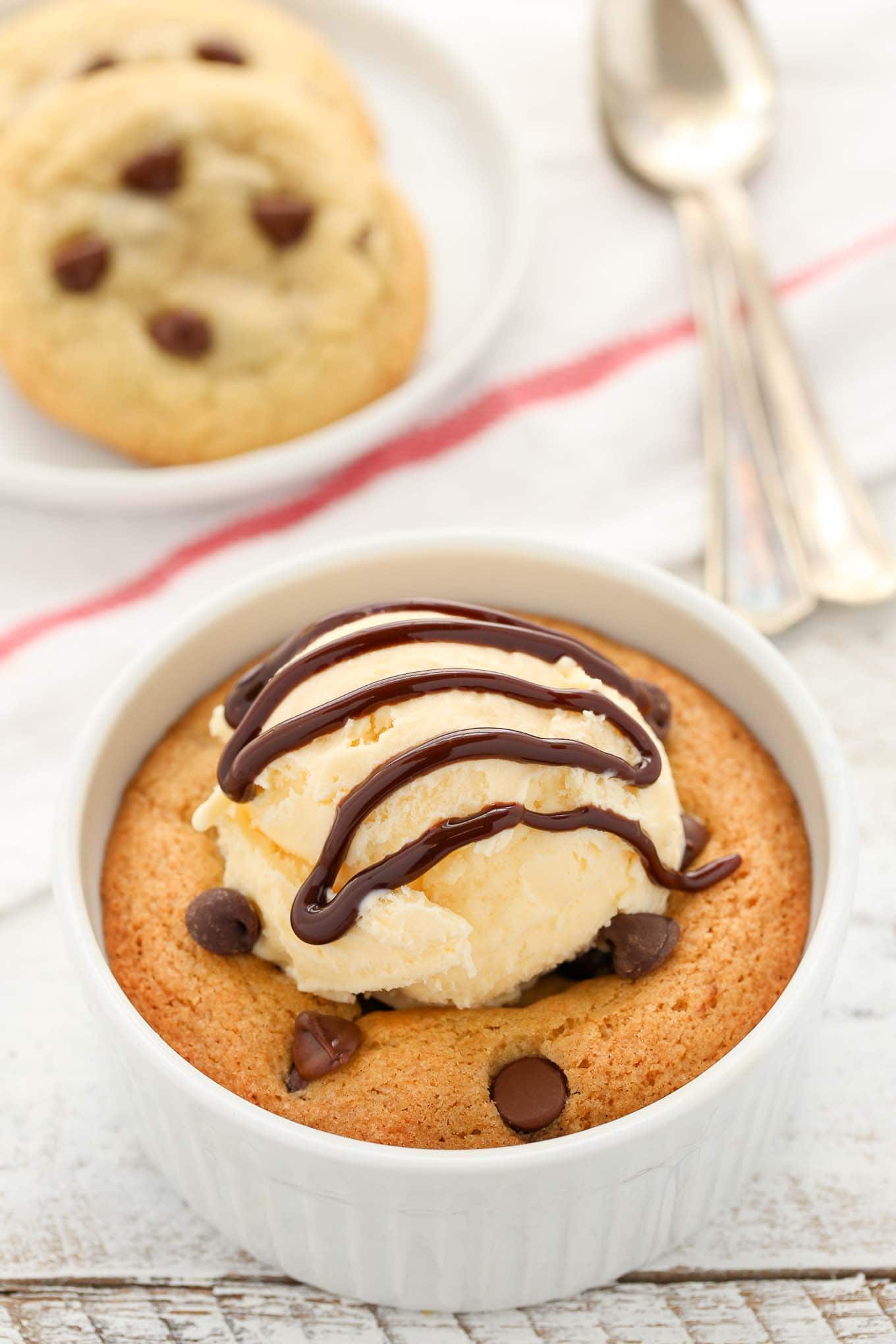 A deep dish single serve chocolate chip cookie topped with ice cream and chocolate sauce. Two cookies rest on a plate in the background. 