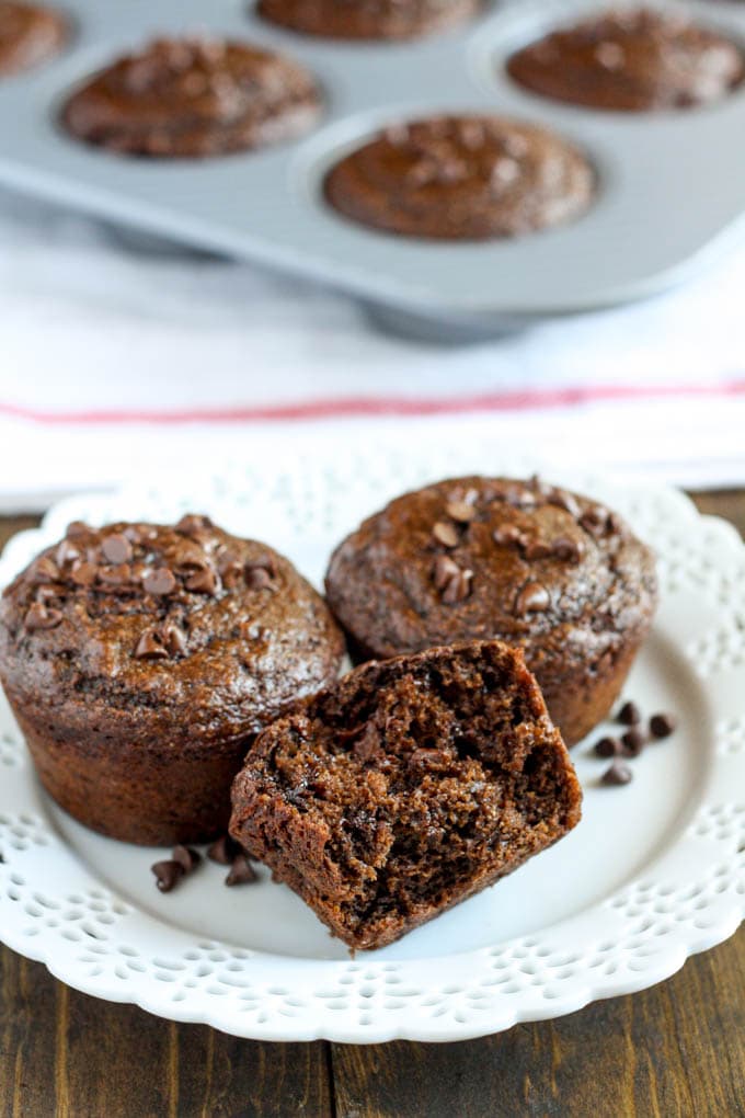 Three healthy chocolate muffins on a white plate. Additional muffins sit in a muffin tin in the background. 