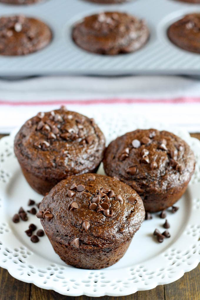 Three healthy chocolate muffins on a white plate. Additional muffins sit in a muffin tin in the background. 