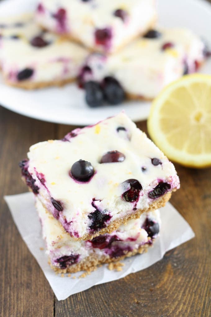 A stack of two lemon blueberry cheesecake bars. A lemon half and additional bars rest in the background. 