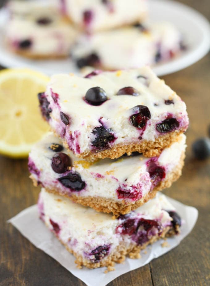 A stack of three lemon blueberry cheesecake bars. A lemon half and additional bars rest in the background. 