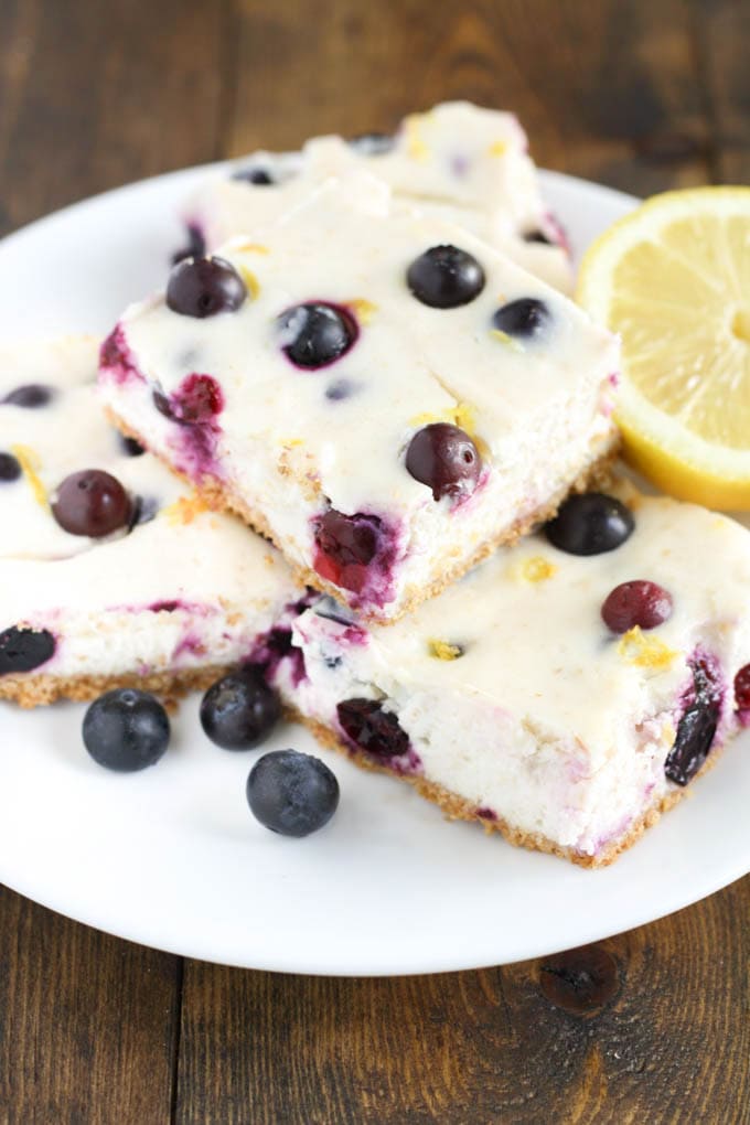 Four lemon blueberry bars piled on a plate, with a lemon half. 